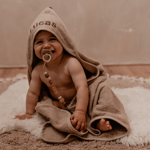 Een newborn baby in een badcape met de naam erop geborduurd en een speenkoord met naam in de mond